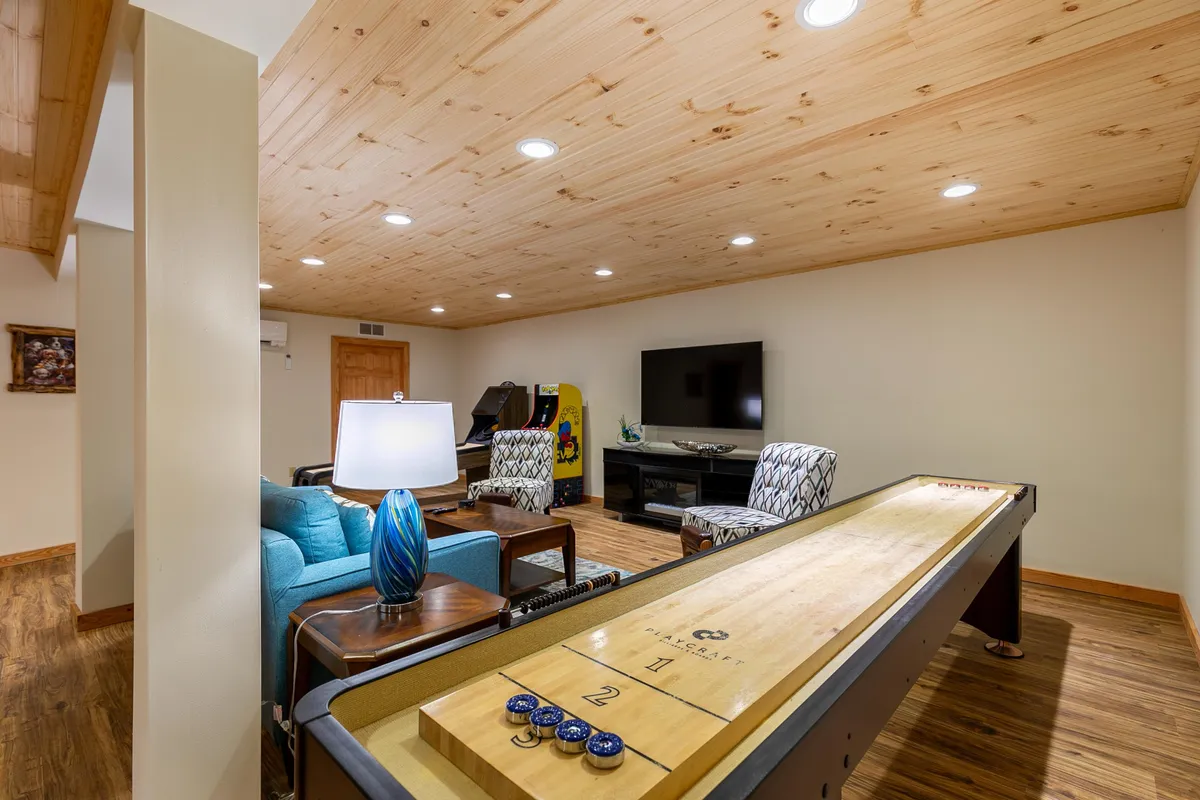 A casual game room setup that includes a shuffleboard table, a blue armchair, a television, and a modern light fixture. The space feels relaxed and is clearly intended for unwinding, friendly competition, and socializing after work.