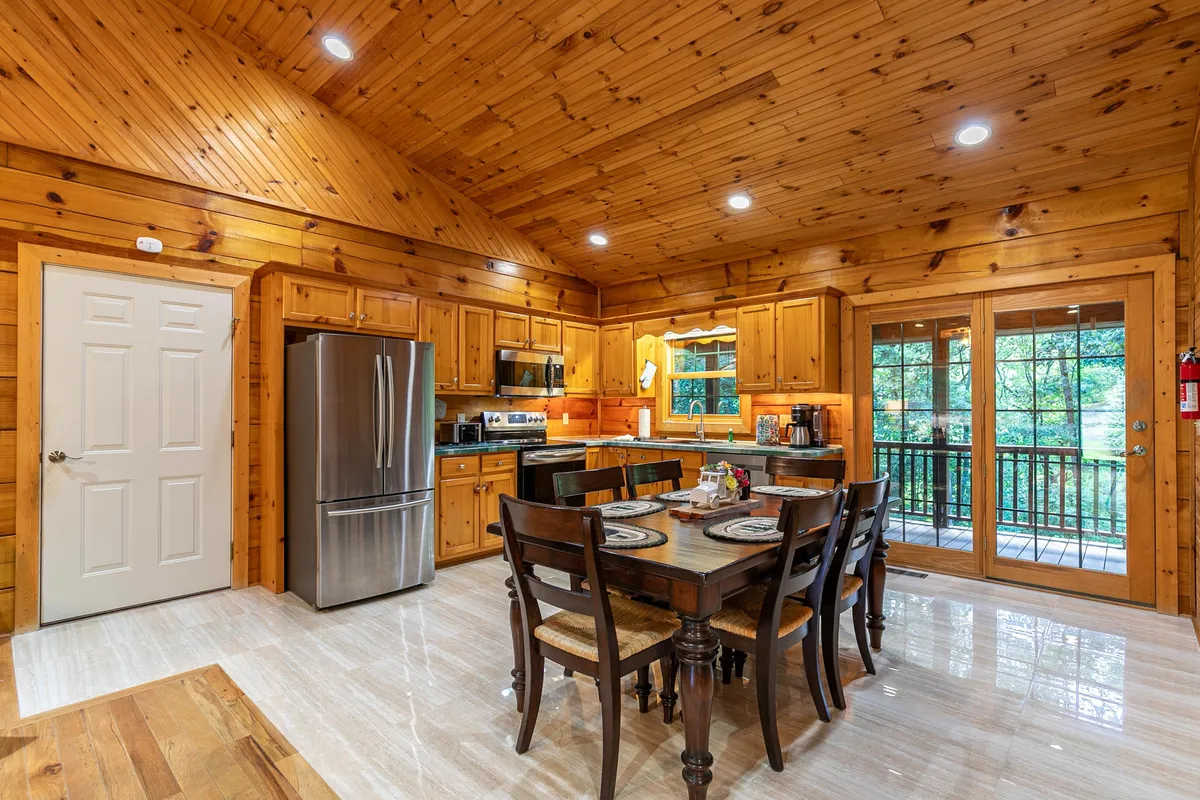 This image captures a warm, welcoming dining area adjacent to the kitchen, featuring a large table perfect for family meals or gatherings. The scene includes visible kitchen appliances, ample wooden cabinetry, and expansive windows that let in natural light and offer a glimpse of the outdoors, reinforcing the open-concept design.