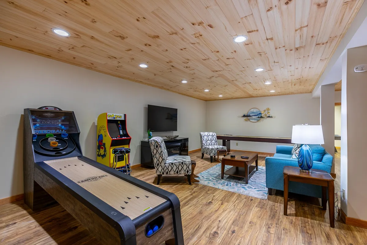 A lively game room featuring a shuffleboard table, an arcade game, a large TV, a comfortable seating area, and decorative wall art. The scene is set for interactive play and casual relaxation, with multiple activities visible at once.