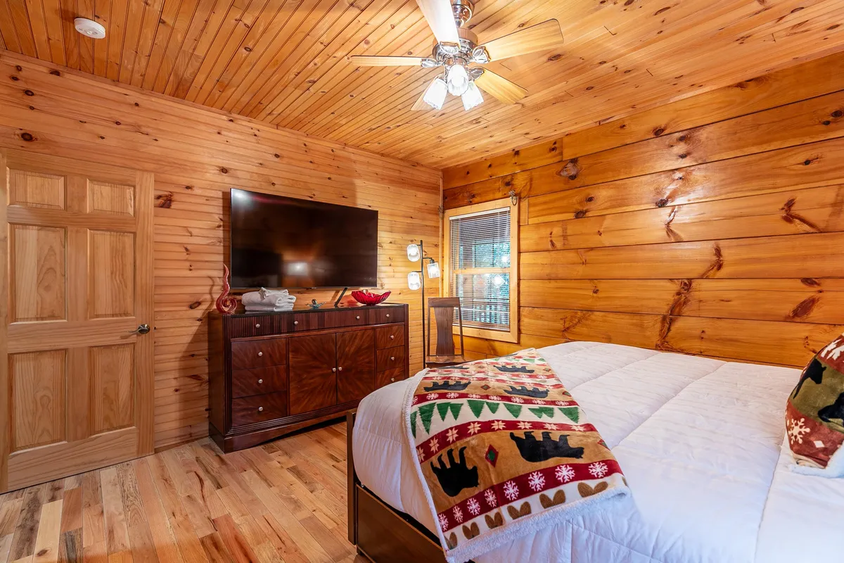 A comfortable sleeping area with a large bed, decorative blanket, and a television, set against warm wooden walls. The space is designed for relaxation and can be linked to late-night lounging or movie marathons.