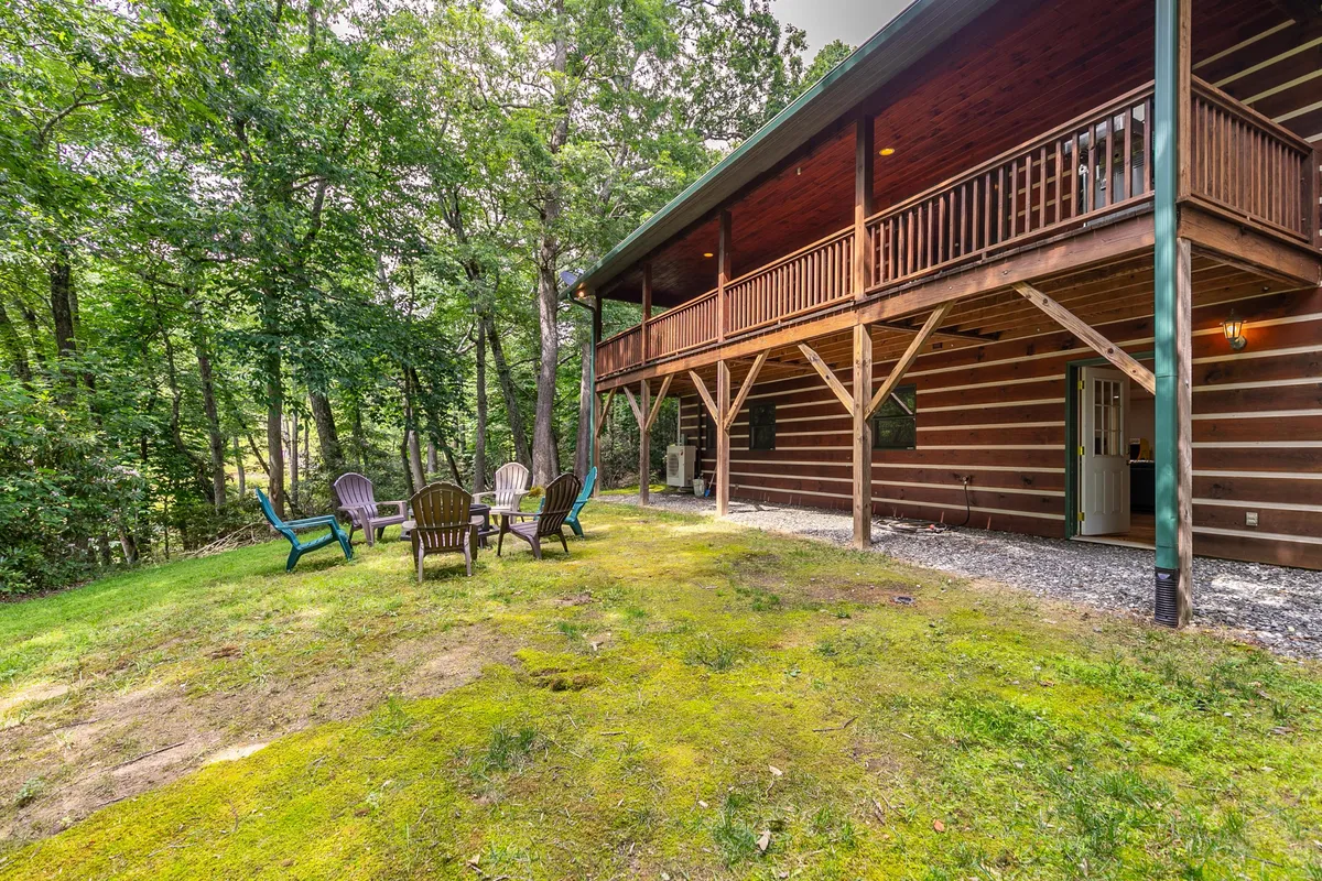 A wooden balcony with comfortable seating overlooks a tranquil wooded area. The scene includes chairs arranged for relaxation and conversation, with mossy ground and tall trees creating a peaceful, inviting outdoor environment.