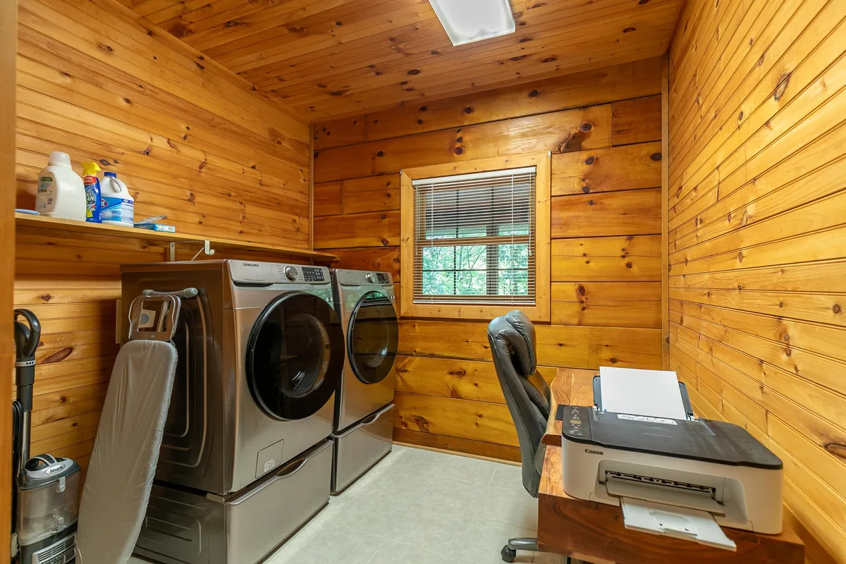 A practical and inviting laundry room featuring a washer and dryer, a printer, wooden walls, and a window that lets in natural light. The room is tidy, with supplies well-organized, and there is a workspace area conducive to remote work tasks.