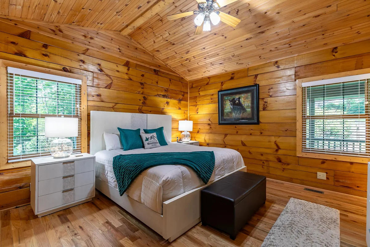 A tranquil bedroom with wooden walls, a bed adorned with decorative pillows, a nightstand featuring a softly glowing lamp, and tasteful artwork on the wall. The warm hues and natural textures create an inviting, peaceful space ideal for rest and relaxation.