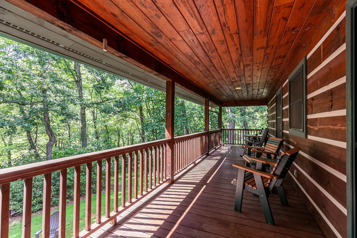 An inviting exterior porch with sturdy wooden chairs facing outwards, surrounded by lush greenery. The wooden railing and natural setting evoke a sense of tranquility and connection to the outdoors.