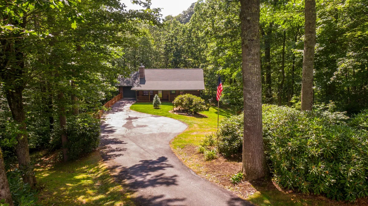 The image showcases a charming log cabin surrounded by a beautifully landscaped yard, a prominent flag pole with an American flag, and inviting outdoor spaces perfect for gatherings. The peaceful setting is enhanced by the soft glow of sunset, promising serene evenings and the opportunity for stargazing from the spacious porch.