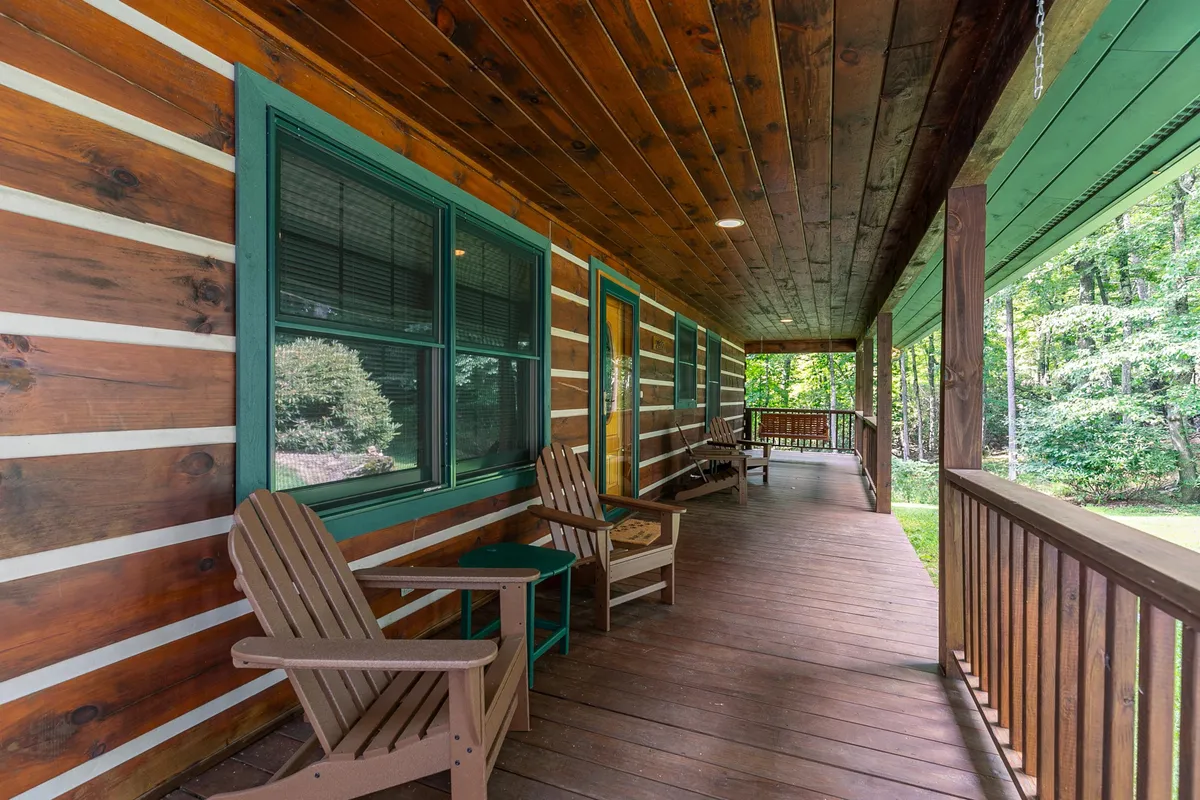 A tranquil exterior shot of the cabin's porch, featuring wooden chairs, log walls, porch railing, and abundant green foliage. The peaceful ambiance encourages relaxation and nature enjoyment.