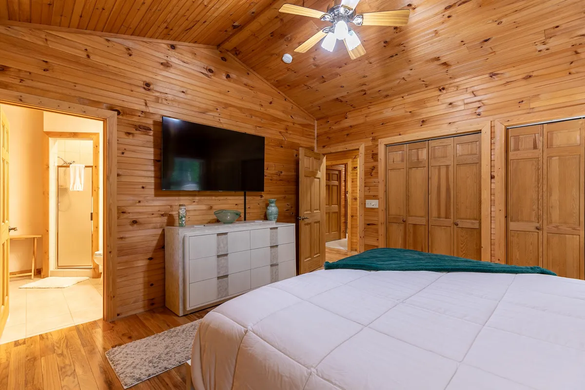 A cozy bedroom featuring wooden walls, a large bed with a crisp white comforter, a dresser with decorative items, and a wall-mounted television. The space feels both modern and relaxing, emphasizing convenience and comfort.