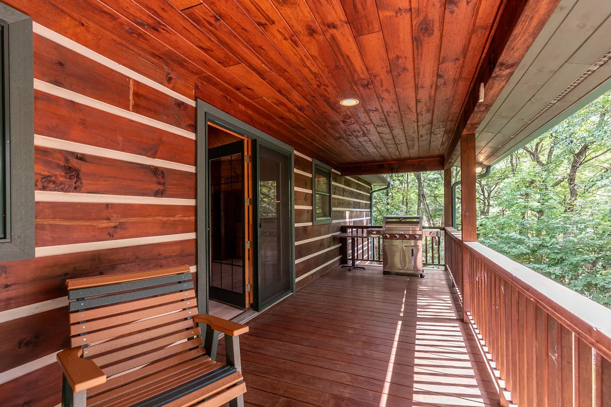 A serene exterior shot showing a wooden chair beside a barbecue grill on a paved patio area, adjacent to the cabin’s exterior wall and sliding glass door, with greenery in the background.