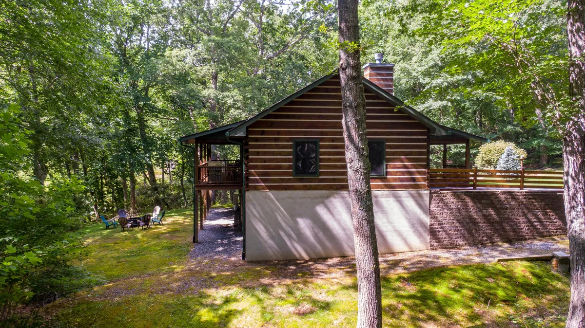 A charming log cabin nestled among tall trees, featuring a spacious outdoor seating area perfect for group gatherings. The porch looks inviting for morning coffee, evening relaxation, or sharing stories around a firepit, with the natural beauty of the woods providing a serene backdrop.