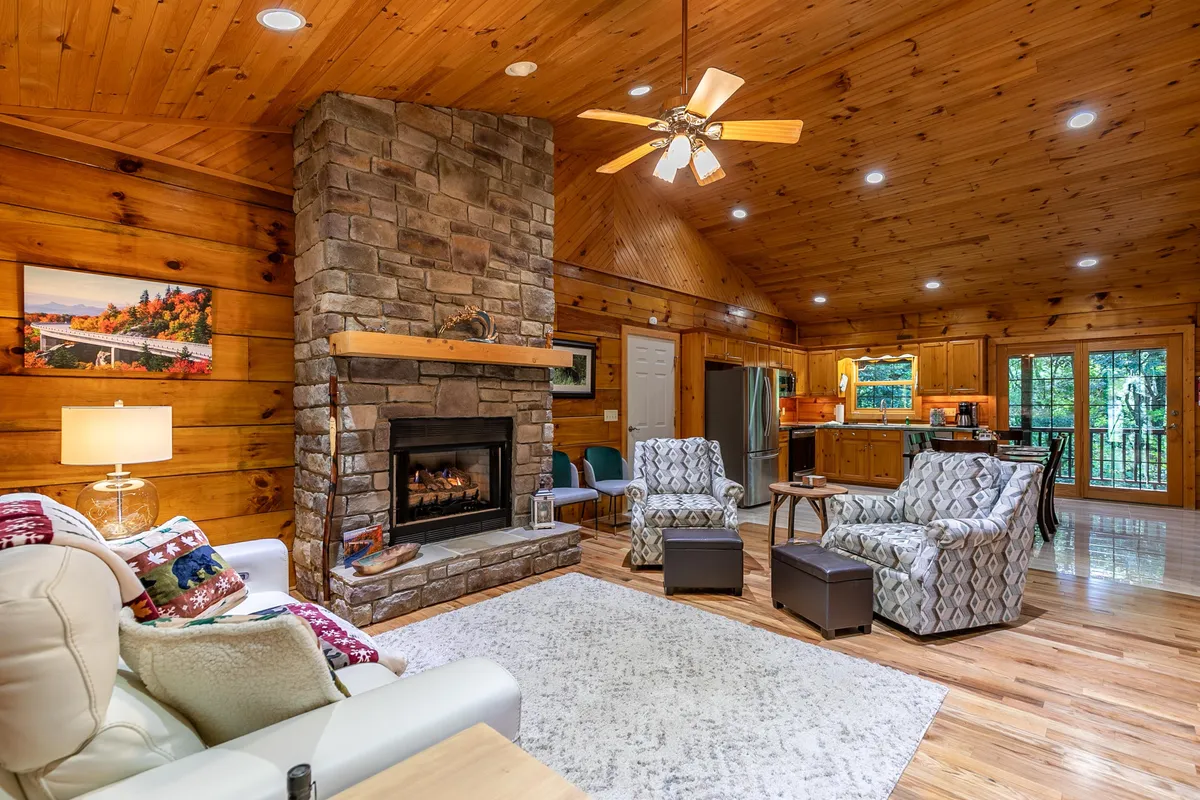A cozy sitting area with a striking stone fireplace, plush seating, and wood-paneled walls. Large windows frame tranquil views of the yard, blending indoor comfort with outdoor serenity and inviting relaxation and conversation.